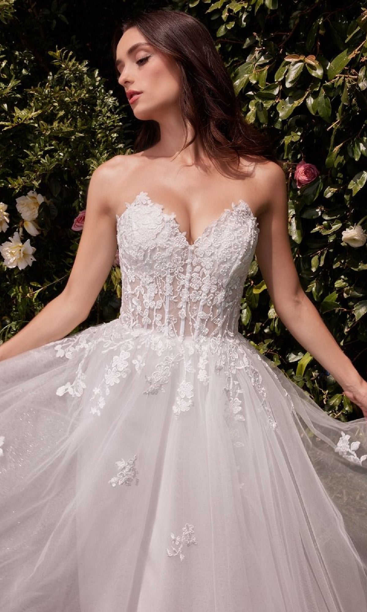 A woman wearing the Strapless Long White Wedding Gown: Ladivine C148W stands outdoors before green foliage and flowers.